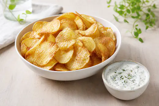 Goudgele zelfgemaakte aardappelchips in een witte kom met een schaaltje yoghurtdip