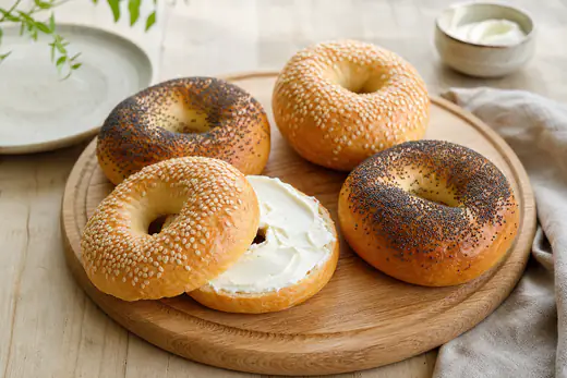 Vier goudbruine bagels met sesamzaad en maanzaad op een houten plank, een ervan doorgesneden met roomkaas