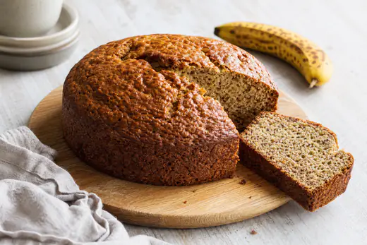 Ronde bananencake met gebarsten goudbruine bovenkant, aangesneden op een houten plank met een plakje ernaast