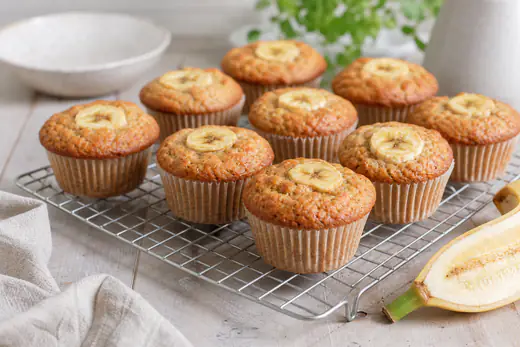 Goudbruine bananenmuffins met een schijfje banaan erop op een rooster, met een halve banaan ernaast