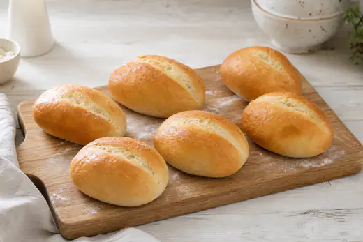 Zes zelfgebakken goudbruine witte broodjes met een ingesneden bovenkant op een houten broodplank