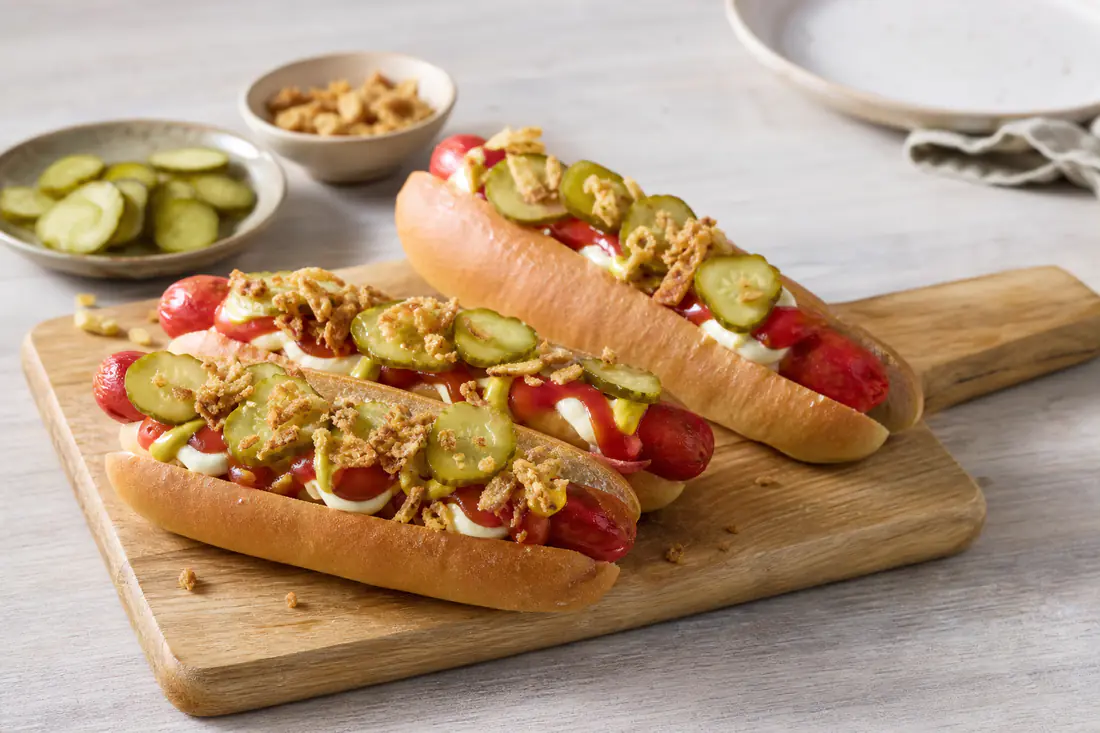 Rode Deense worstjes in hotdogbroodjes met remoulade, gebakken uitjes en plakjes augurk op een houten plank