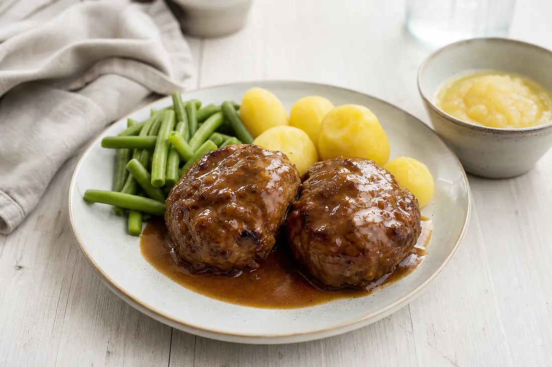 Twee grote bruingebakken gehaktballen met jus, gekookte aardappelen en sperziebonen op een bord