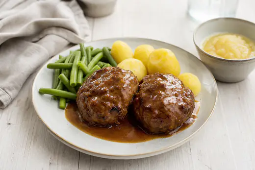 Twee grote bruingebakken gehaktballen met jus, gekookte aardappelen en sperziebonen op een bord
