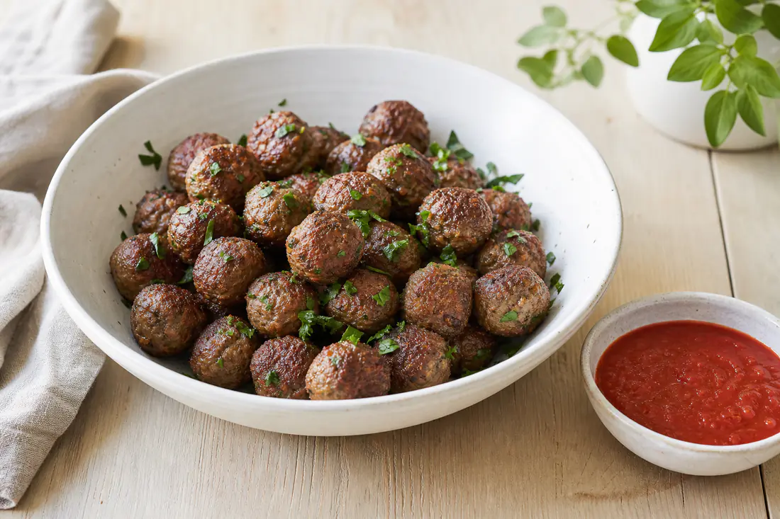 Kleine bruingebakken gehaktballetjes in een witte schaal met gehakte peterselie en een bakje tomatensaus