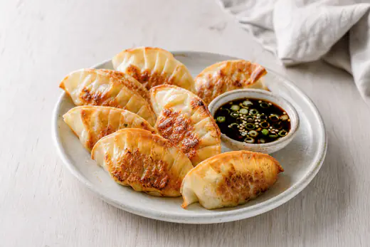 Gouden, halvemaanvormige gyoza met geplisseerde rand op een bord naast een bakje dipsaus met lente-ui en sesamzaad