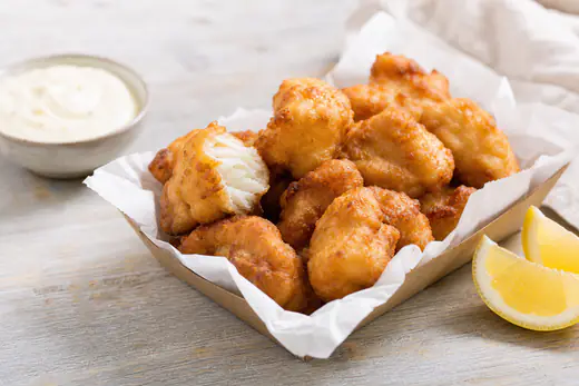 Goudbruine stukjes kibbeling, gepaneerde kabeljauw, in een papieren bakje met een schaaltje knoflooksaus en een partje citroen