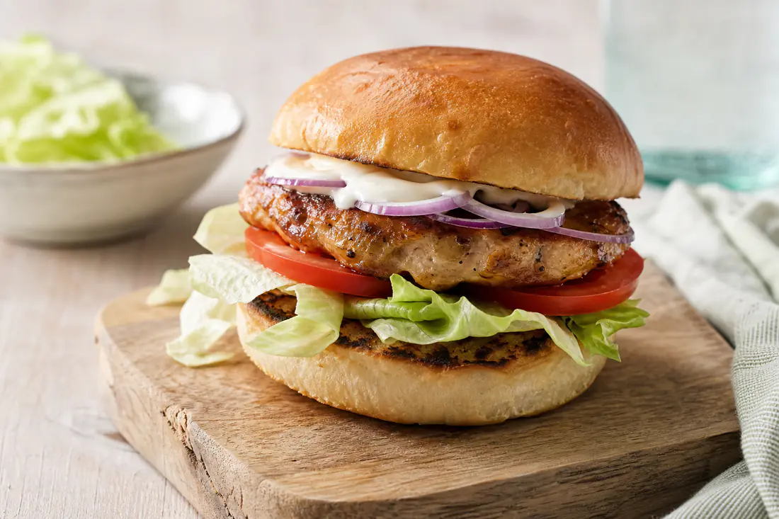 Kipburger op een geroosterd broodje met ijsbergsla, tomaat, rode ui en mayonaise op een plank