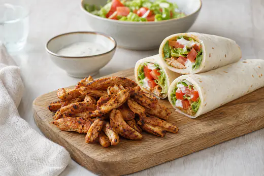 Gekruide kipreepjes met paprika en komijn op een plank naast opgerolde wraps met sla en knoflooksaus