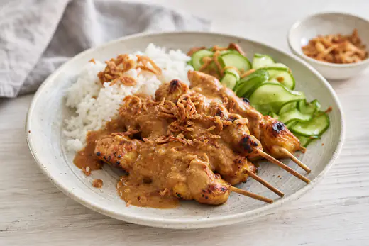 Satéstokjes met gegrilde kip en pindasaus op een bord met witte rijst, komkommer en gebakken uitjes