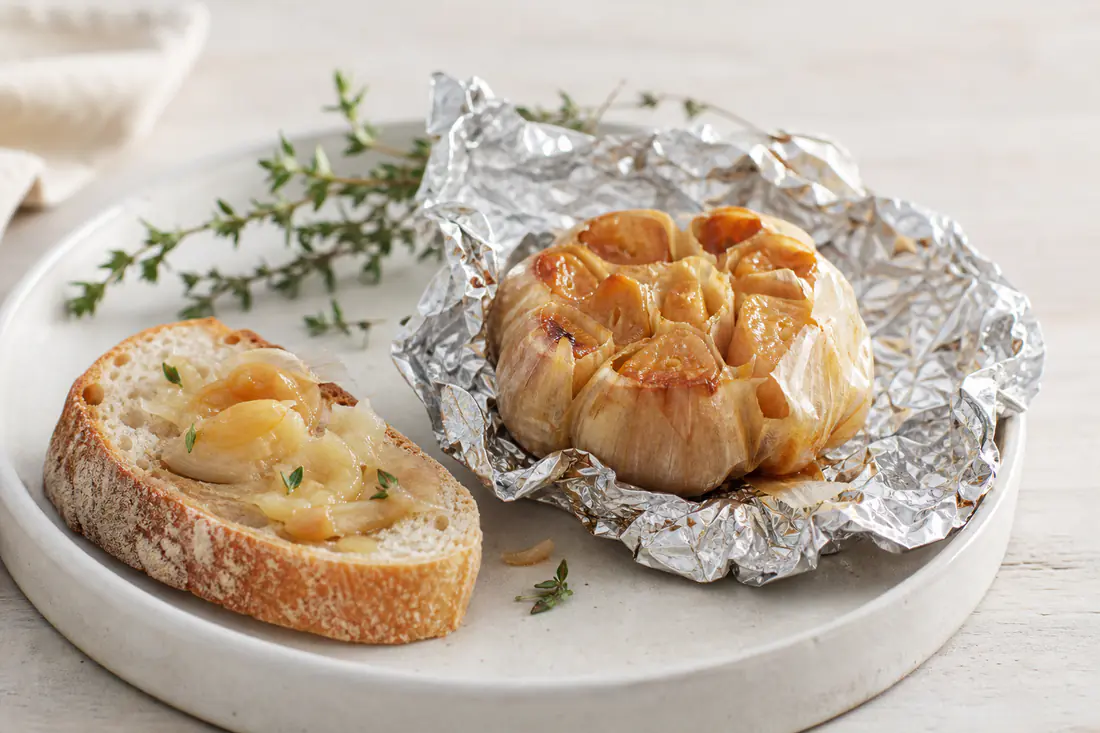 Gepofte knoflookbol in aluminiumfolie, opengesneden met zachte gouden teentjes en een stuk brood