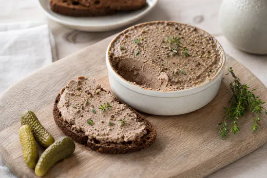 Zelfgemaakte leverpastei in een klein ovenschaaltje met een plak op roggebrood, augurkjes en verse tijm
