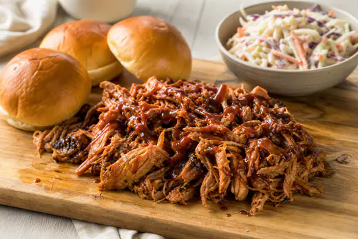 Uit elkaar getrokken pulled pork met barbecuesaus op een houten plank, met zachte broodjes en koolsalade ernaast