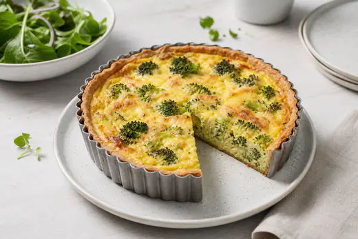 Kleine ronde quiche met broccoli en kaas in een bakvorm, een punt eruit gesneden met stevige gele vulling en gouden korst