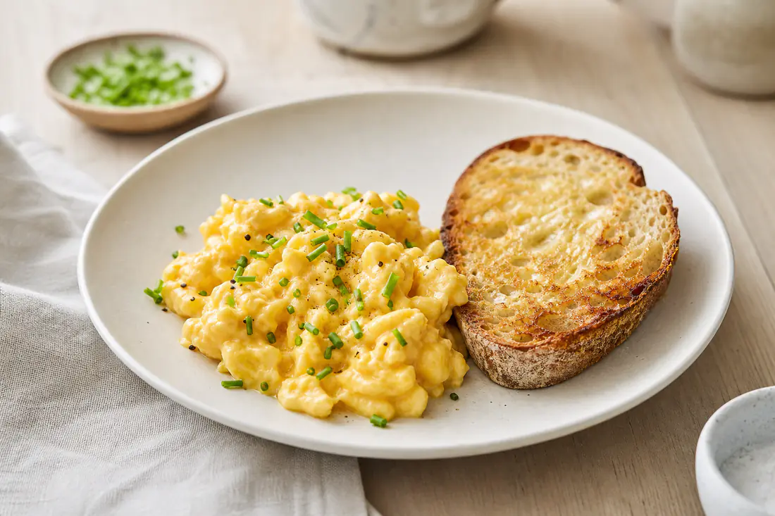 Romig roerei met bieslook op een bord naast een snee geroosterd brood