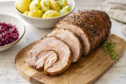 Gebakken varkensrollade in plakken gesneden met een kruidenkorst en een sappig roze-bruin snijvlak naast gekookte aardappelen