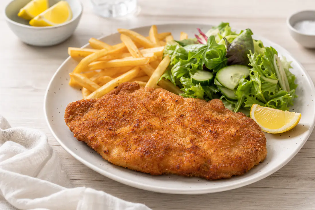 Goudbruine gepaneerde varkensschnitzel op een bord met een partje citroen, friet en een frisse salade