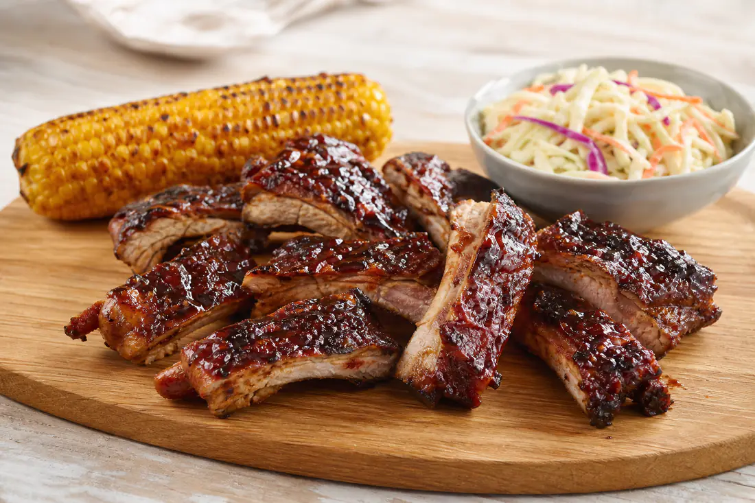 Glanzende spareribs met barbecueglaze, in stukken gesneden op een houten plank met maiskolf en coleslaw