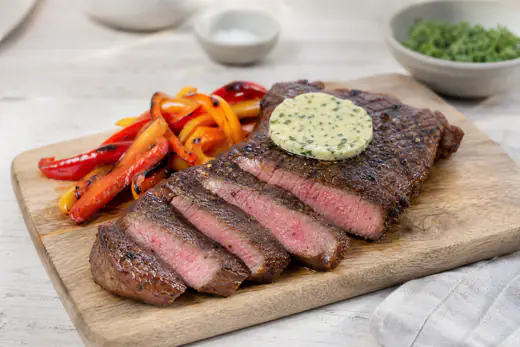 Aangesneden biefstuk met roze binnenkant en een plakje kruidenboter op een plank met geroosterde paprika