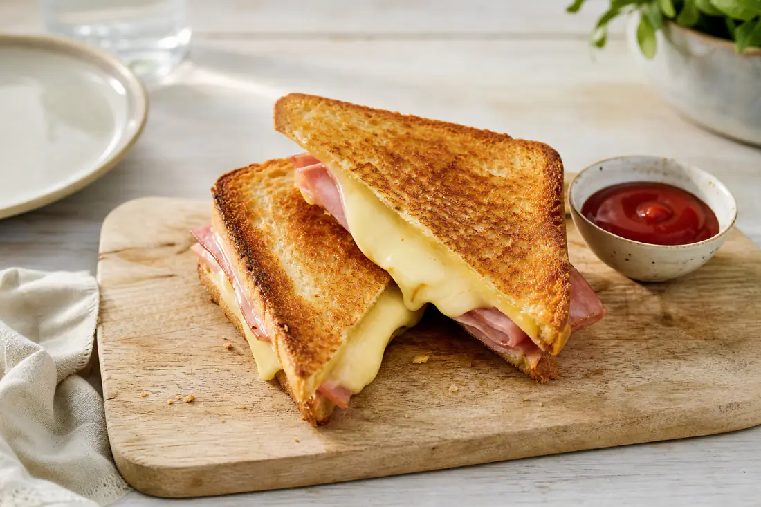 Goudbruine tosti ham-kaas diagonaal doorgesneden met uitlopende gesmolten kaas op een snijplank met ketchup