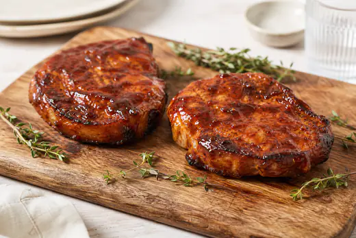 Twee goudbruine varkensribeyes met een glanzend laagje barbecuesaus op een houten plank met tijm