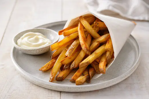 Zelfgesneden goudbruine friet met schilrandjes in een papieren puntzak met een bakje mayonaise ernaast